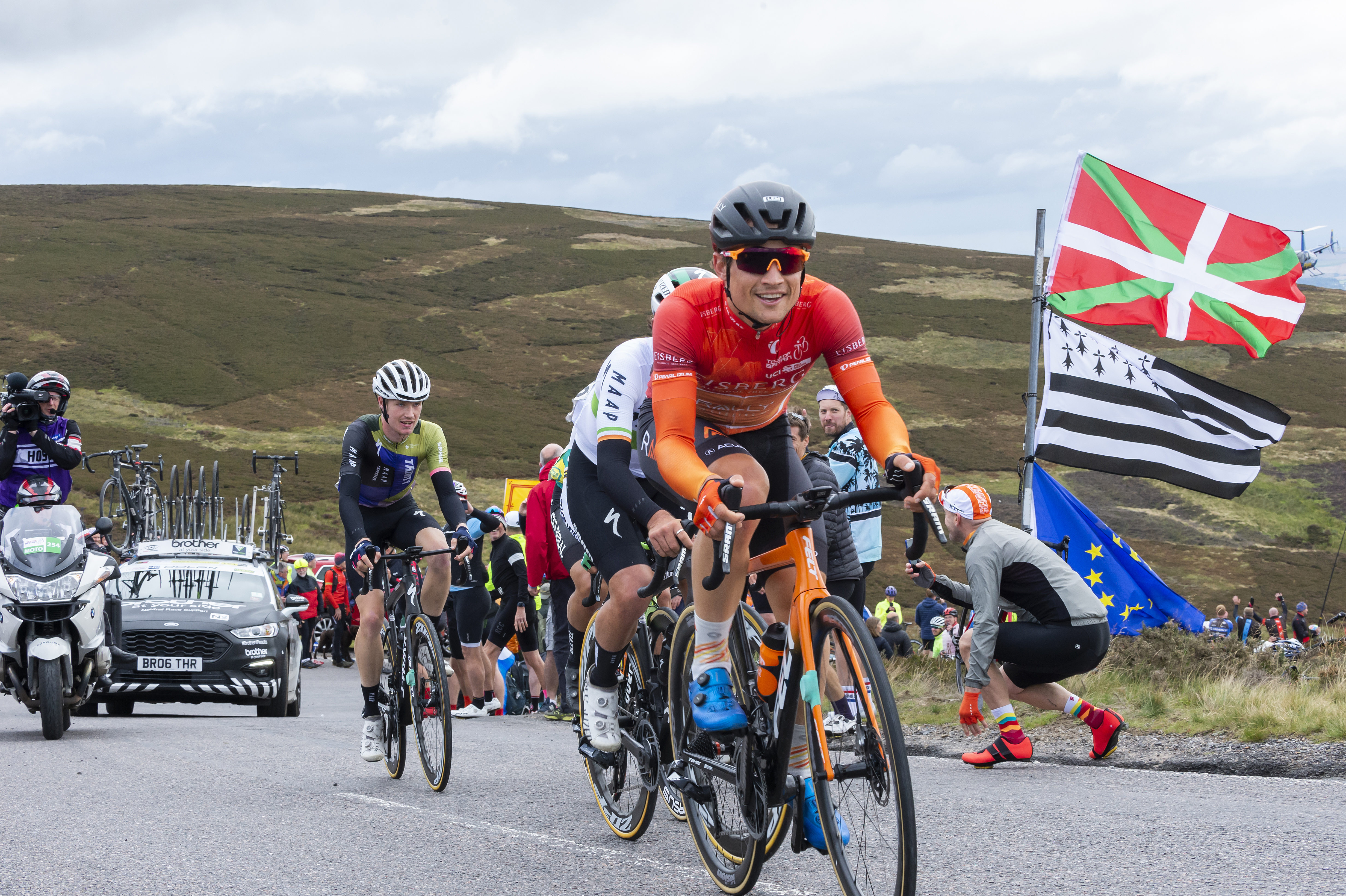 Riders cycling up a hill in Aberdeenshire with support cars and flags in the background.